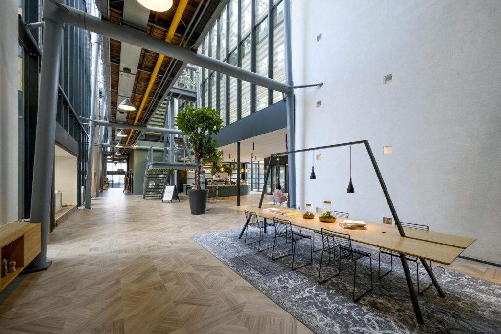 Moderne kantoorlobby aan de H.J.E. Wenckebachweg met hoge plafonds, glazen gevel, en een lange houten vergadertafel onder hanglampen.